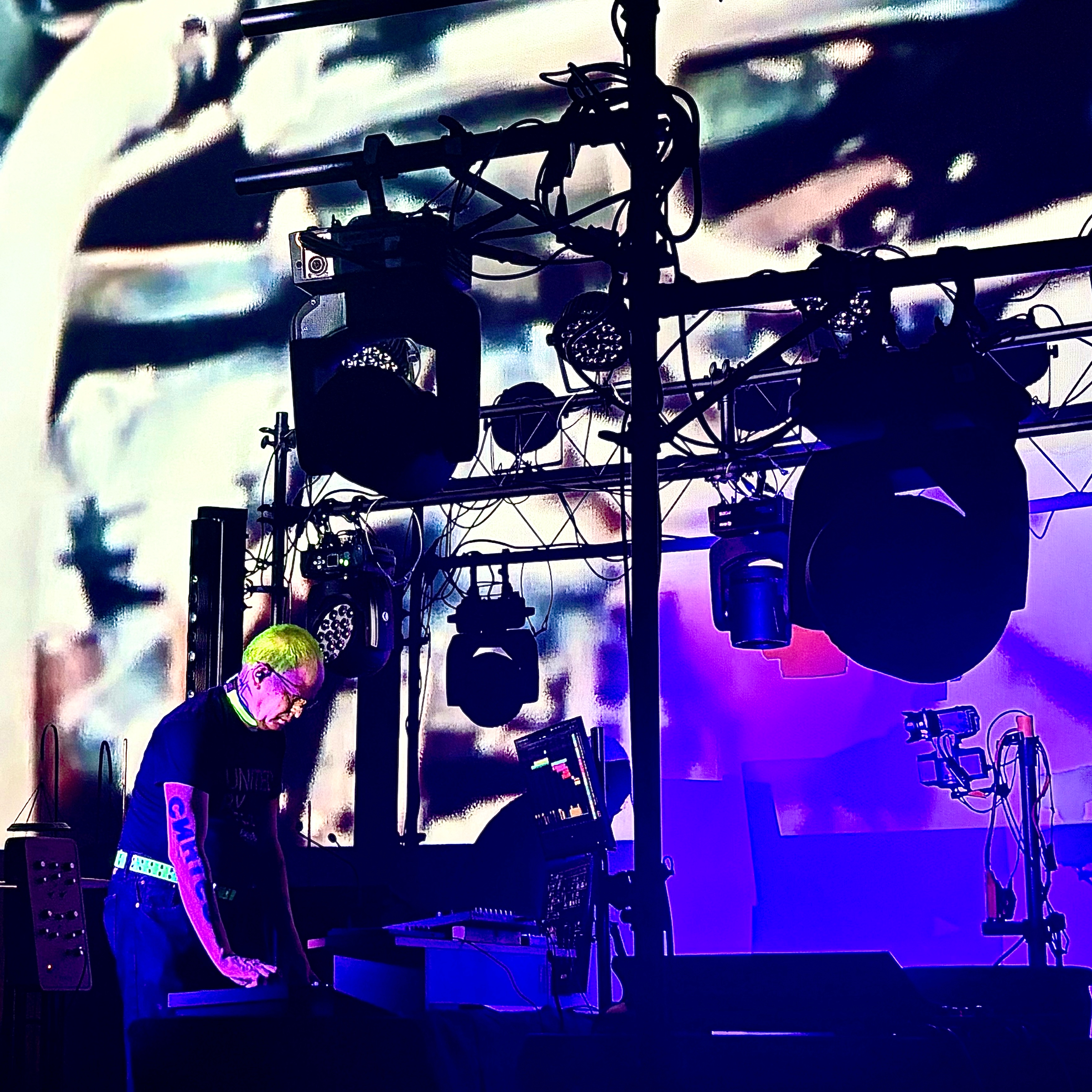 Bill Nobes with neon-green hair leaning over gear on a stage lit in purple and blue